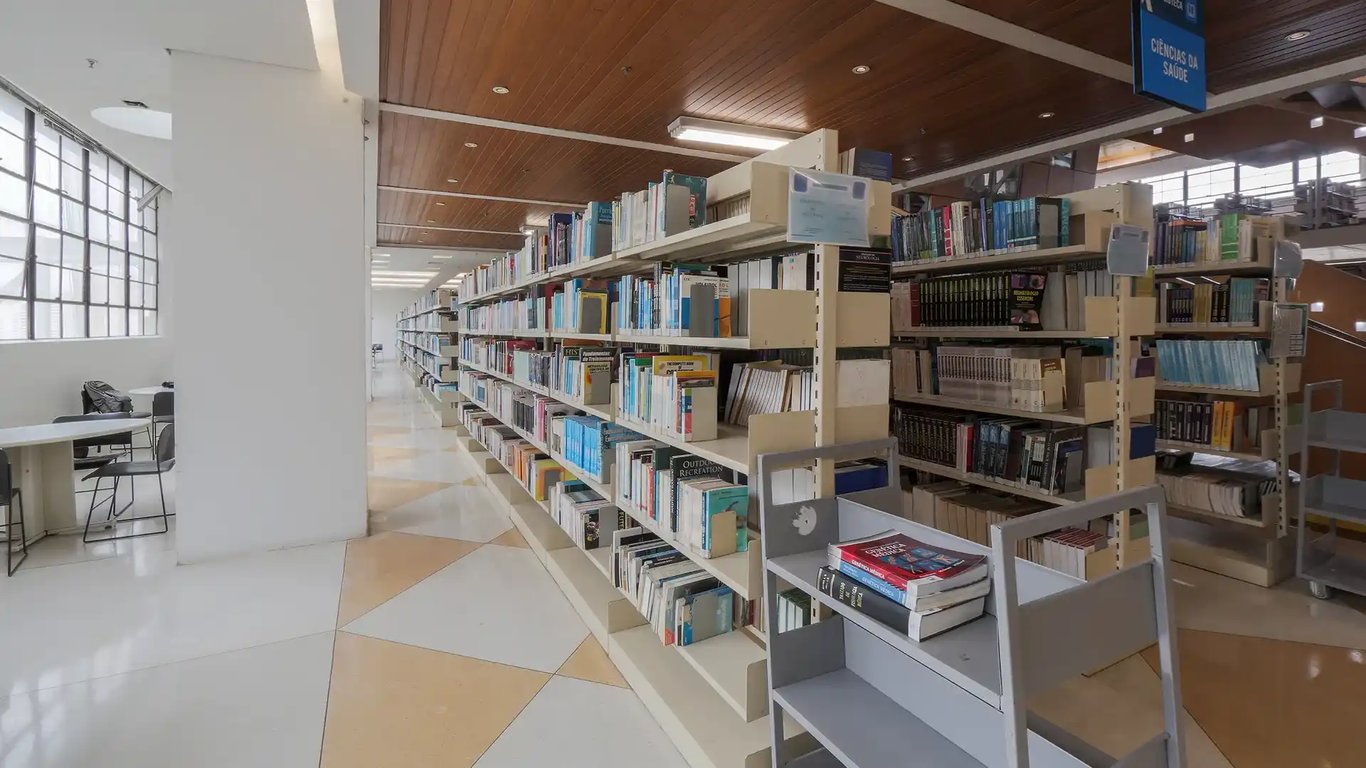 Foto do interior de uma biblioteca moderna com estantes cheias de livros e mesas para estudo. O ambiente é claro e bem organizado, indicando um espaço acadêmico.