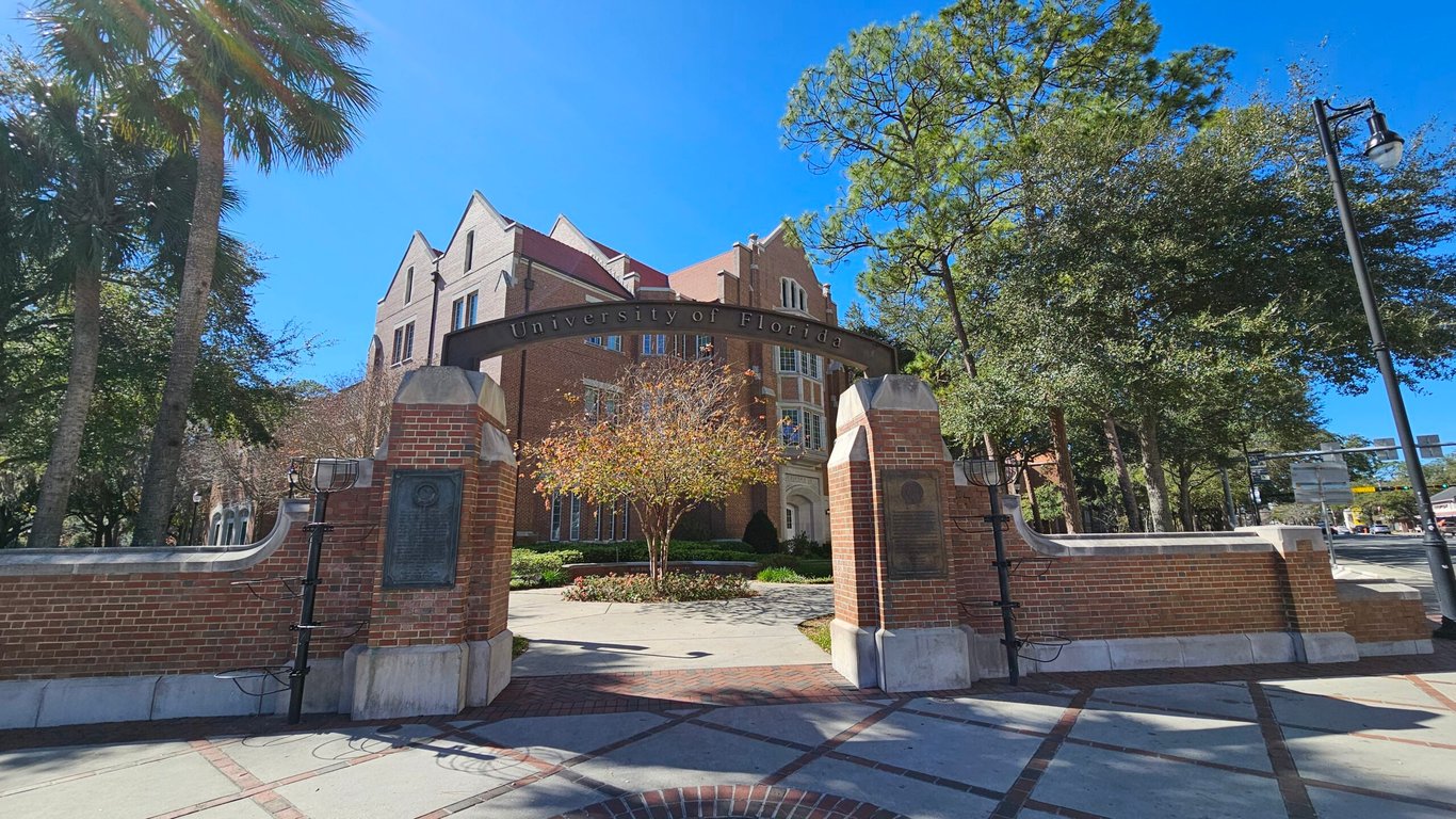 Entrada de campus universitário com vegetação e arquitetura moderna, estudantes caminhando