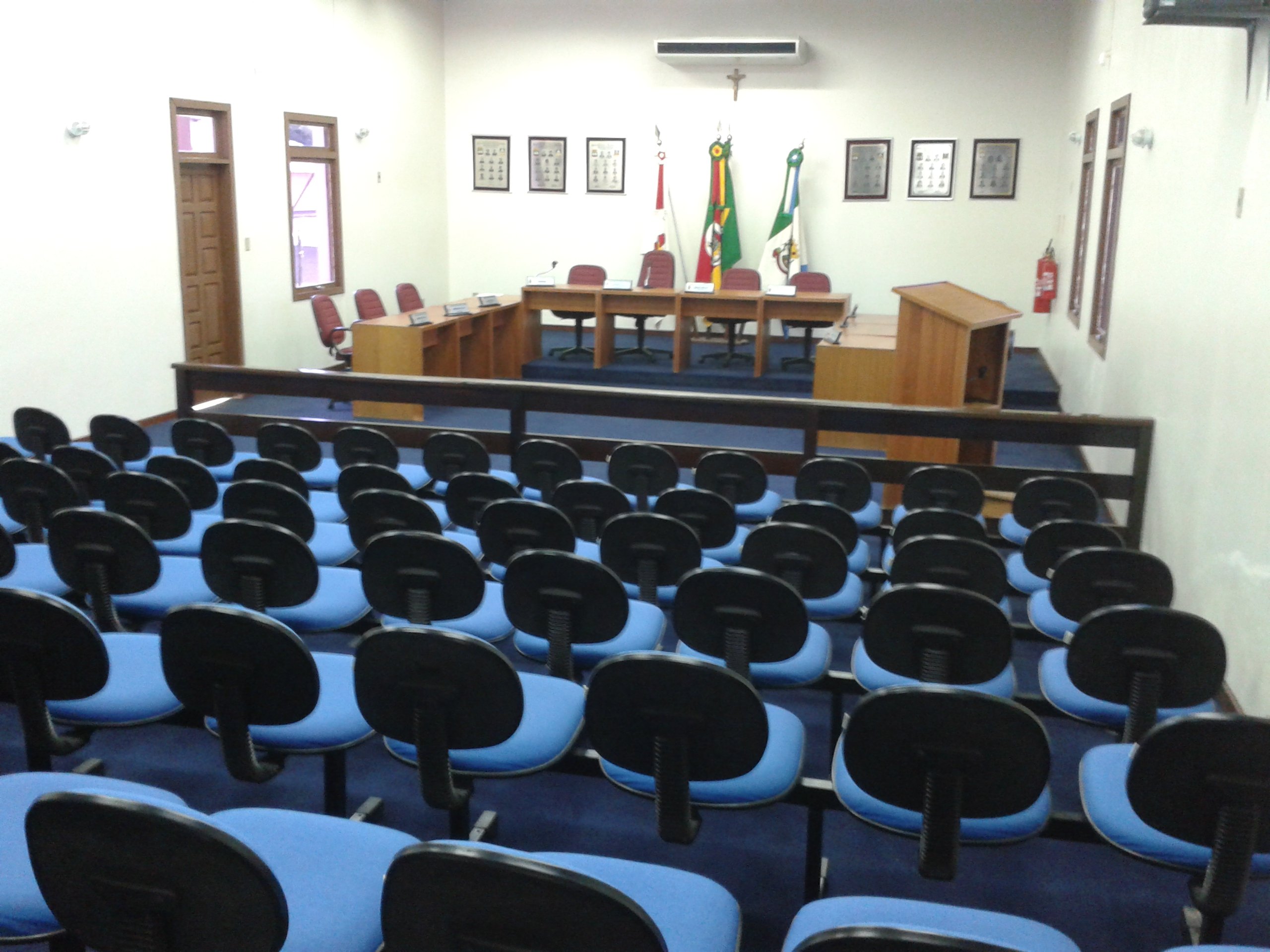 Ambiente interno de um plenário de Câmara Municipal, com mesa diretora e bandeiras
