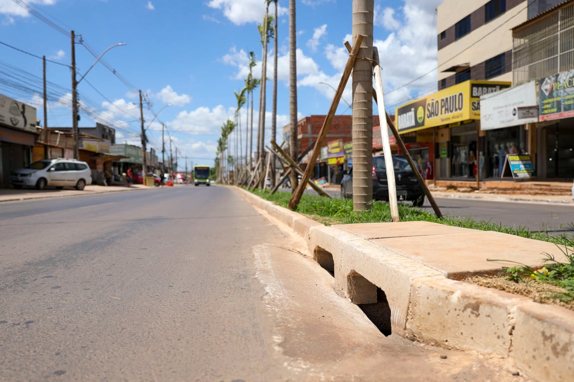 Paisagem urbana de cidade pequena do Centro-Oeste, com via asfaltada e comércio de bairro, sob céu azul com nuvens leves