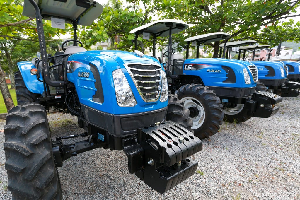 Tratores agrícolas alinhados em área rural, sob luz suave, com paisagem que remete a vinhedos