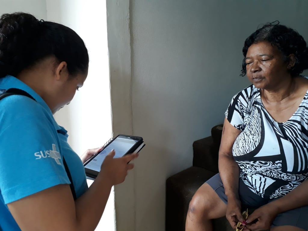 Agente Comunitário de Saúde em atendimento domiciliar, utilizando tablet e conversando com família