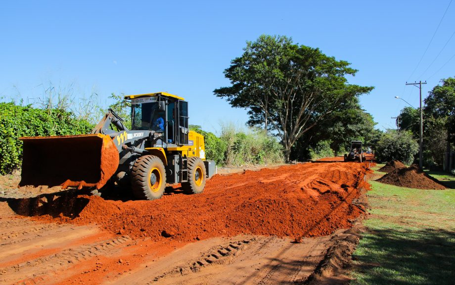 Estrada vicinal de terra sendo trabalhada por máquina ao entardecer