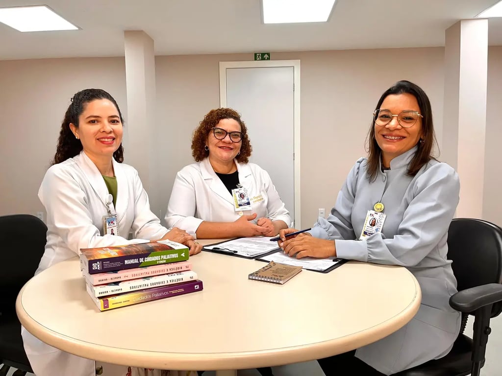 Foto de profissionais de saúde em ambiente ambulatorial. Três mulheres em jalecos brancos sentadas em uma mesa com livros e documentos.