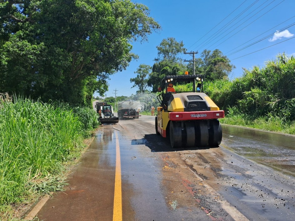 Manutenção de estrada rural com motoniveladora em operação; colinas verdes e céu nublado ao fundo