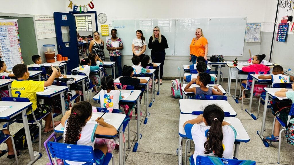 Sala de aula de escola pública com professoras e alunos em atividade, ambiente iluminado, quadro branco ao fundo