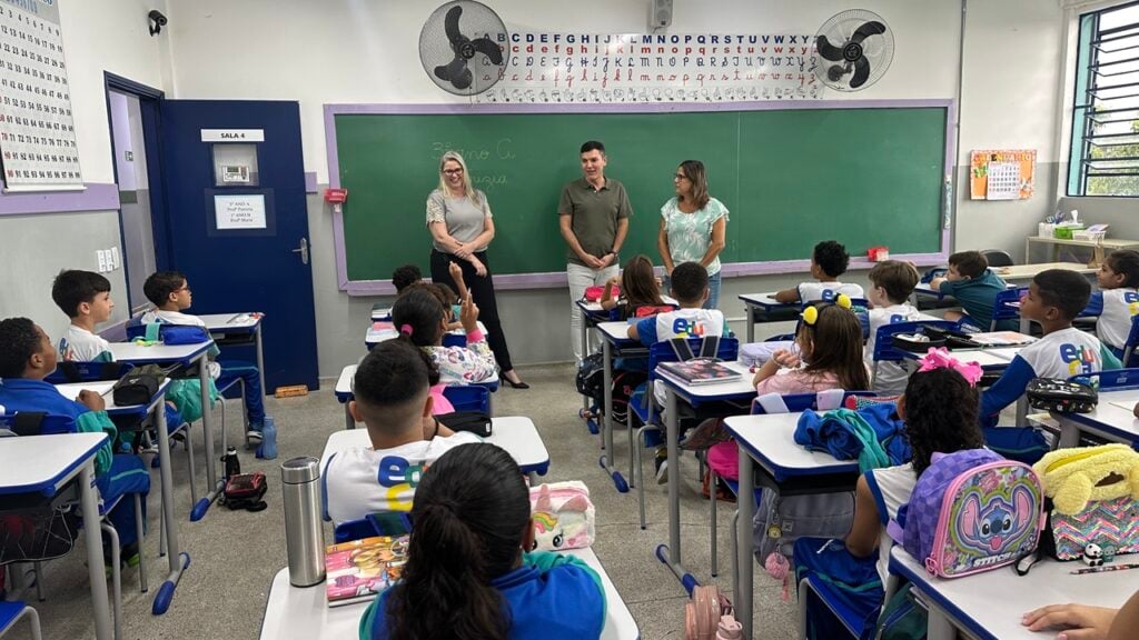 Sala de aula em escola municipal brasileira, com carteiras e lousa, alunos desfocados e iluminação natural
