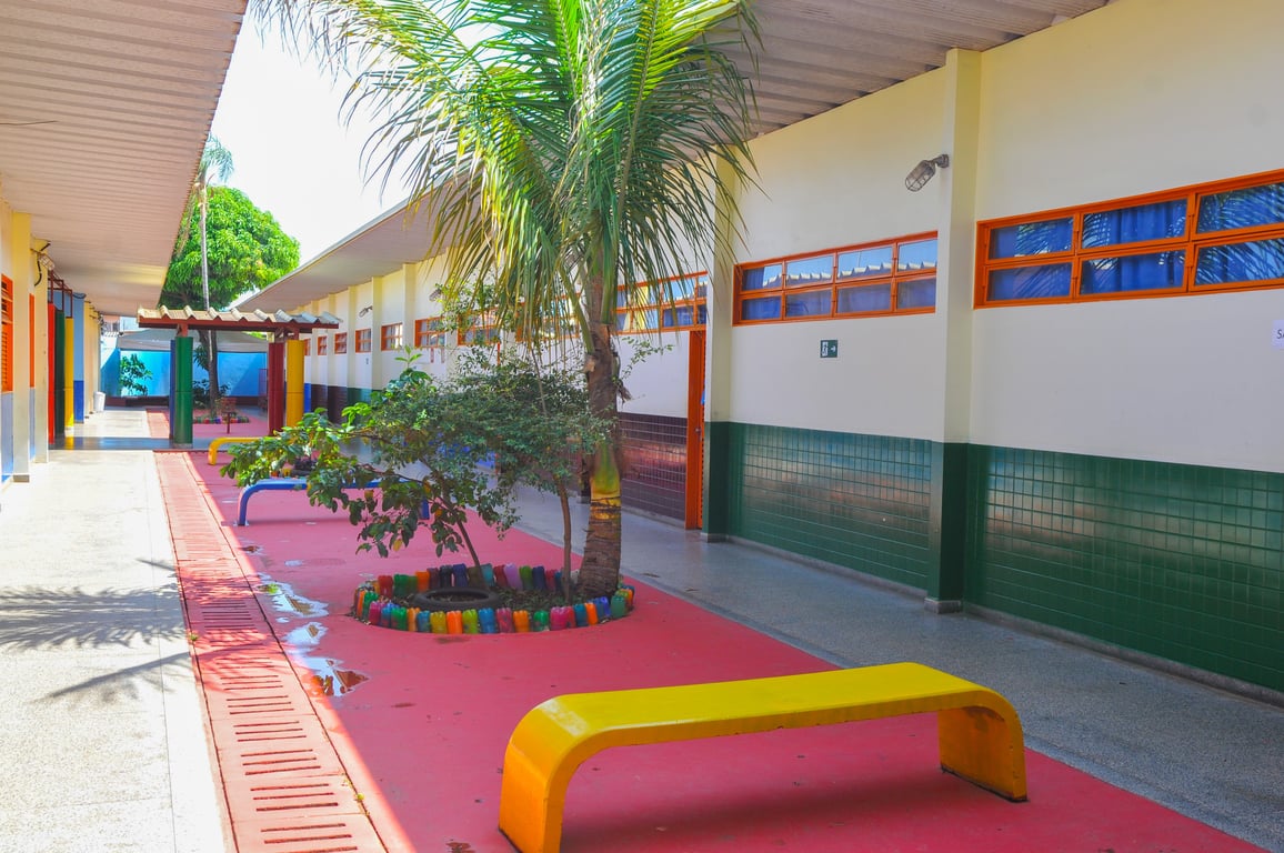 Pátio de escola pública com bancos coloridos sob árvores, em dia ensolarado