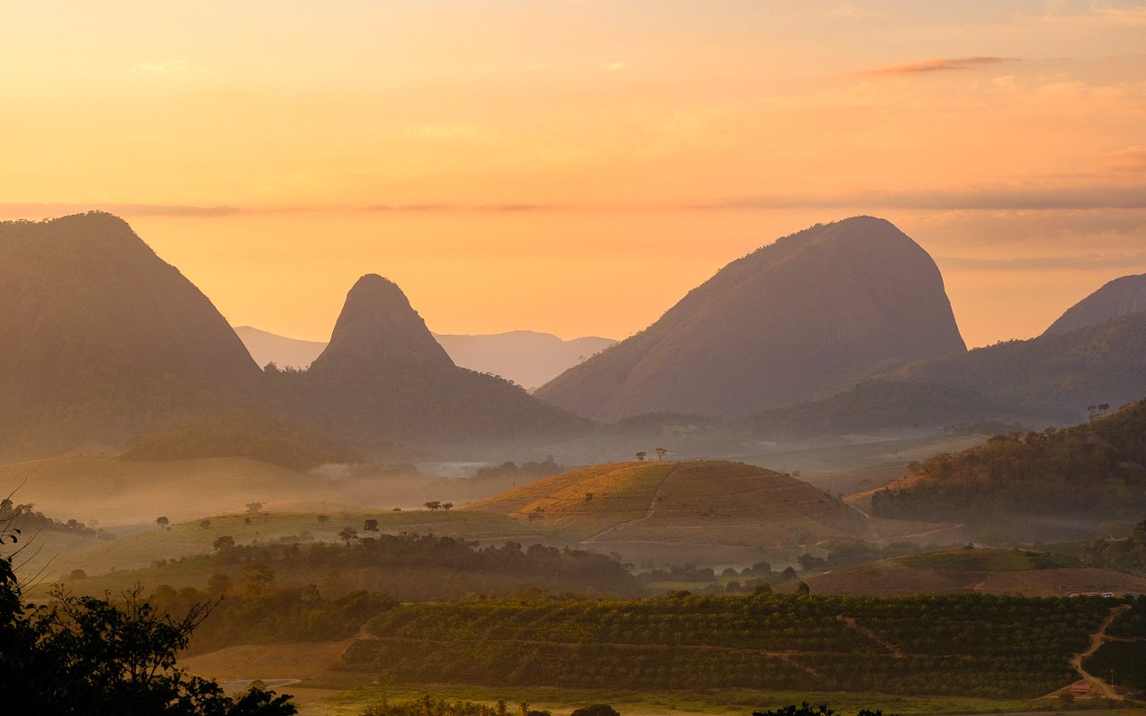 Paisagem de montanhas e vales na região serrana do Espírito Santo ao entardecer, com vegetação atlântica e áreas rurais.