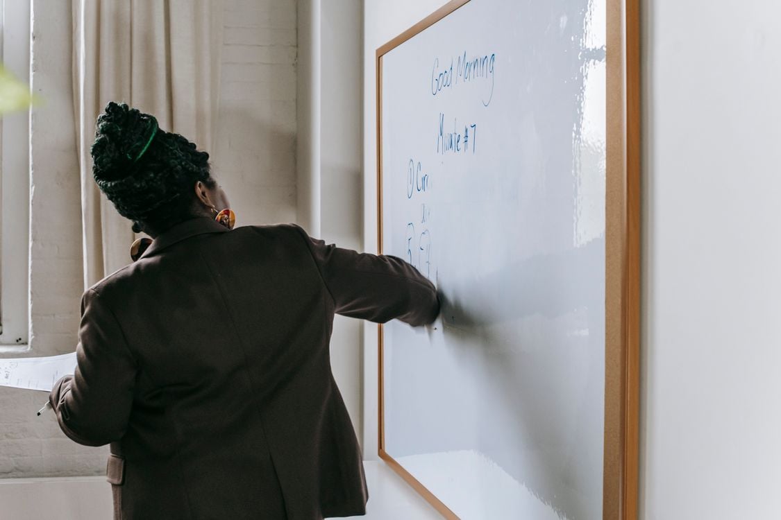 Professora escrevendo no quadro com alunos desfocados ao fundo