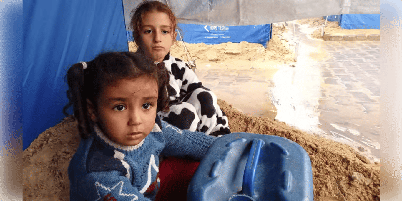 Two young girls sitting beside a water container in a sandy tent camp
