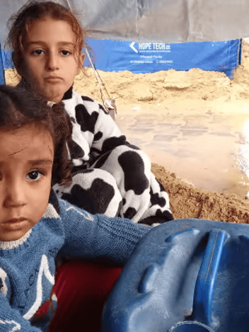 Two young girls sitting beside a water container in a sandy tent camp