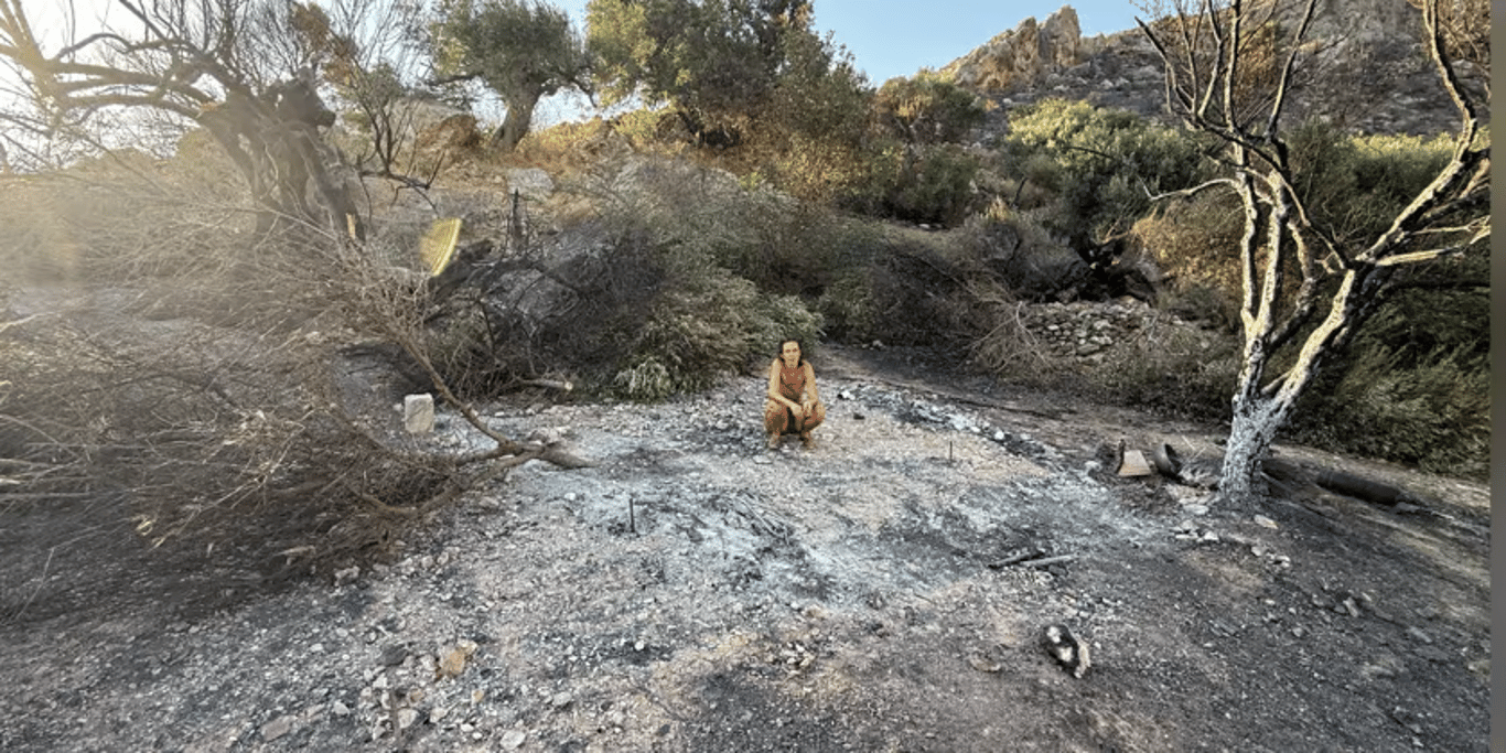 A woman crouching on scorched ground among burnt trees after a wildfire