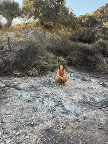 A woman crouching on scorched ground among burnt trees after a wildfire