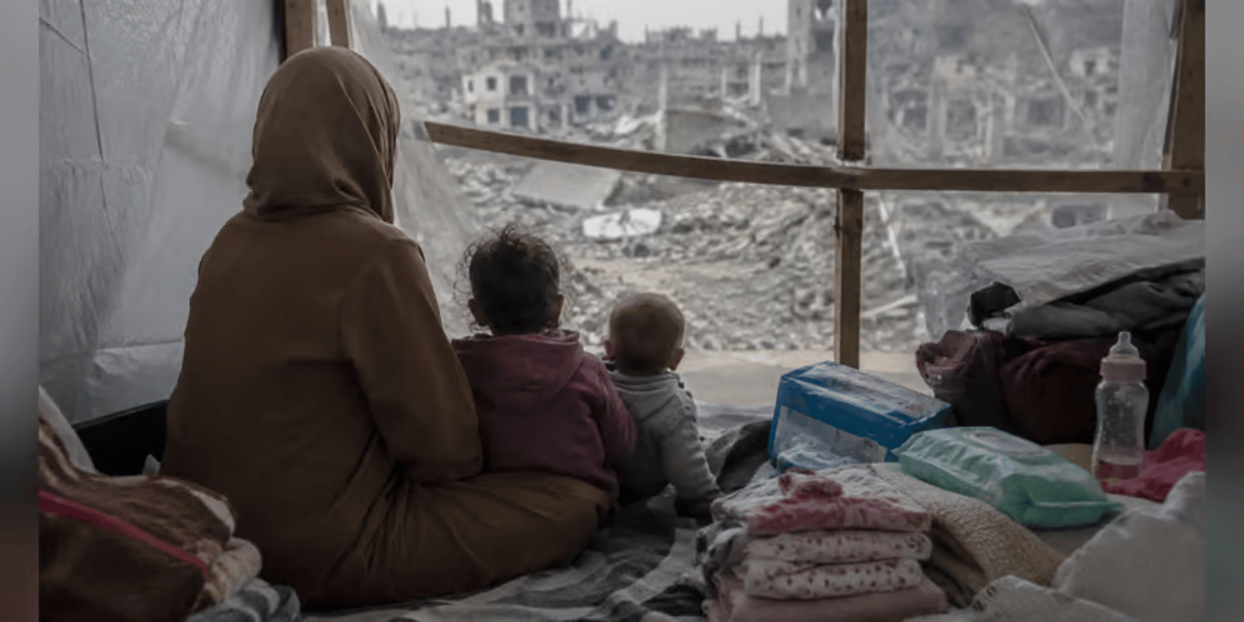 A mother and two small children sitting in a tent, looking out over destroyed buildings