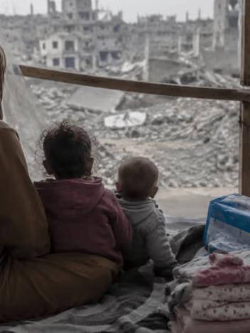 A mother and two small children sitting in a tent, looking out over destroyed buildings