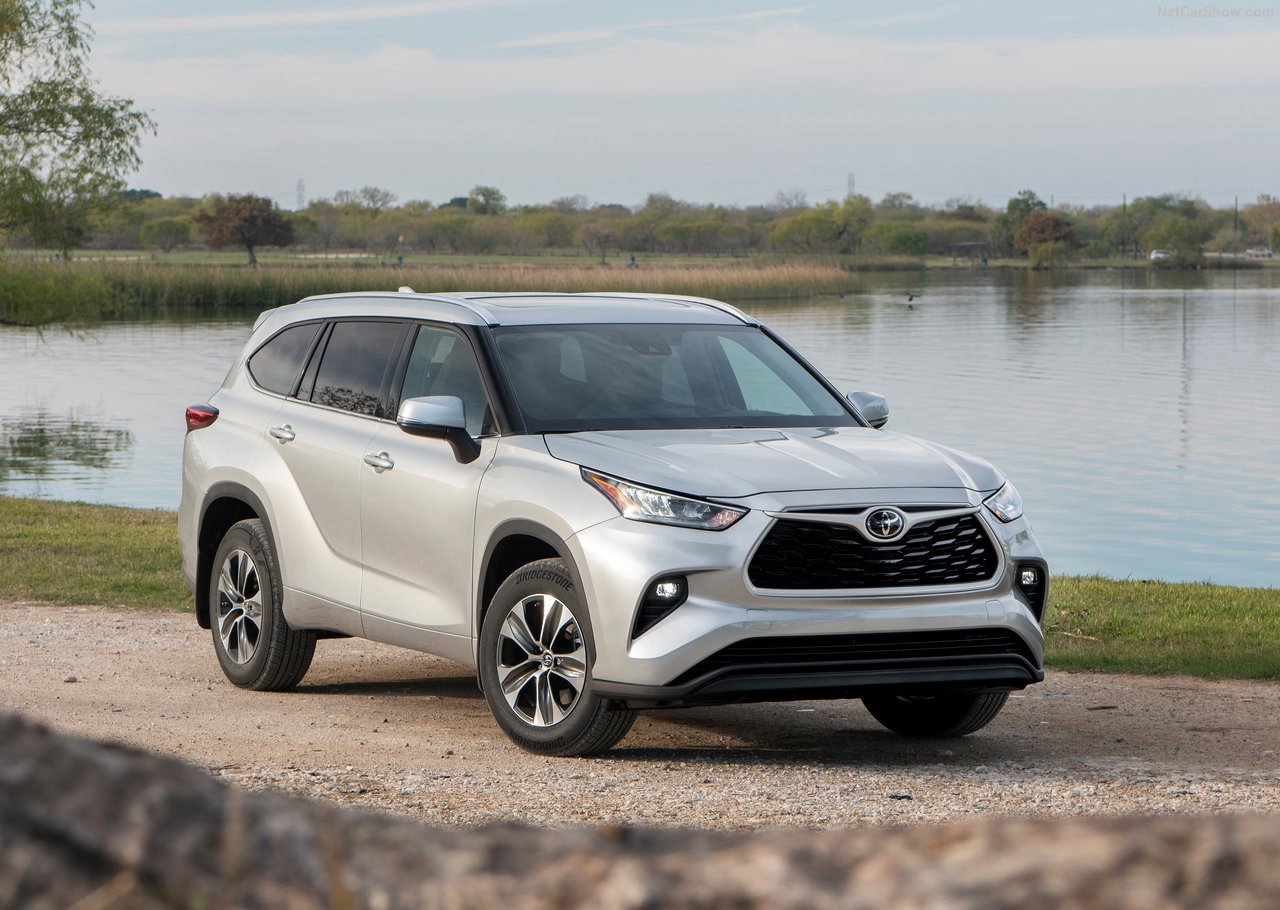 A silver Toyota Highlander, a refined 7 seater car, parked on a gravel path by a tranquil lakeside with trees and clear skies in the background
