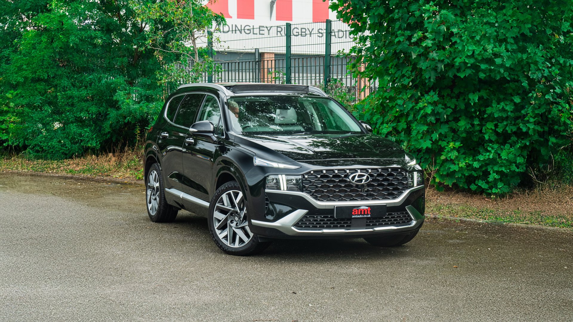 A black Hyundai Santa Fe, a modern 7 seater car, parked on a smooth tarmac surface with a backdrop of vibrant green trees