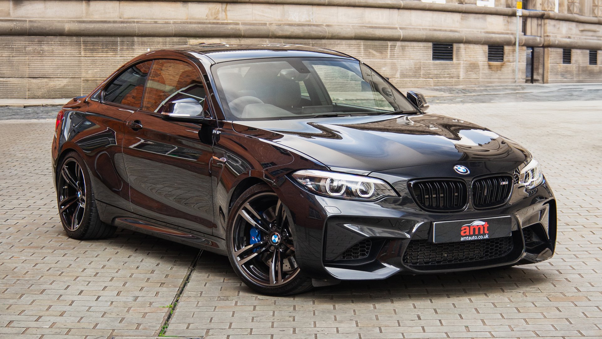 A black BMW M2 captured in an urban setting, parked on a tiled street with its sharp lines and sporty stance emphasised.