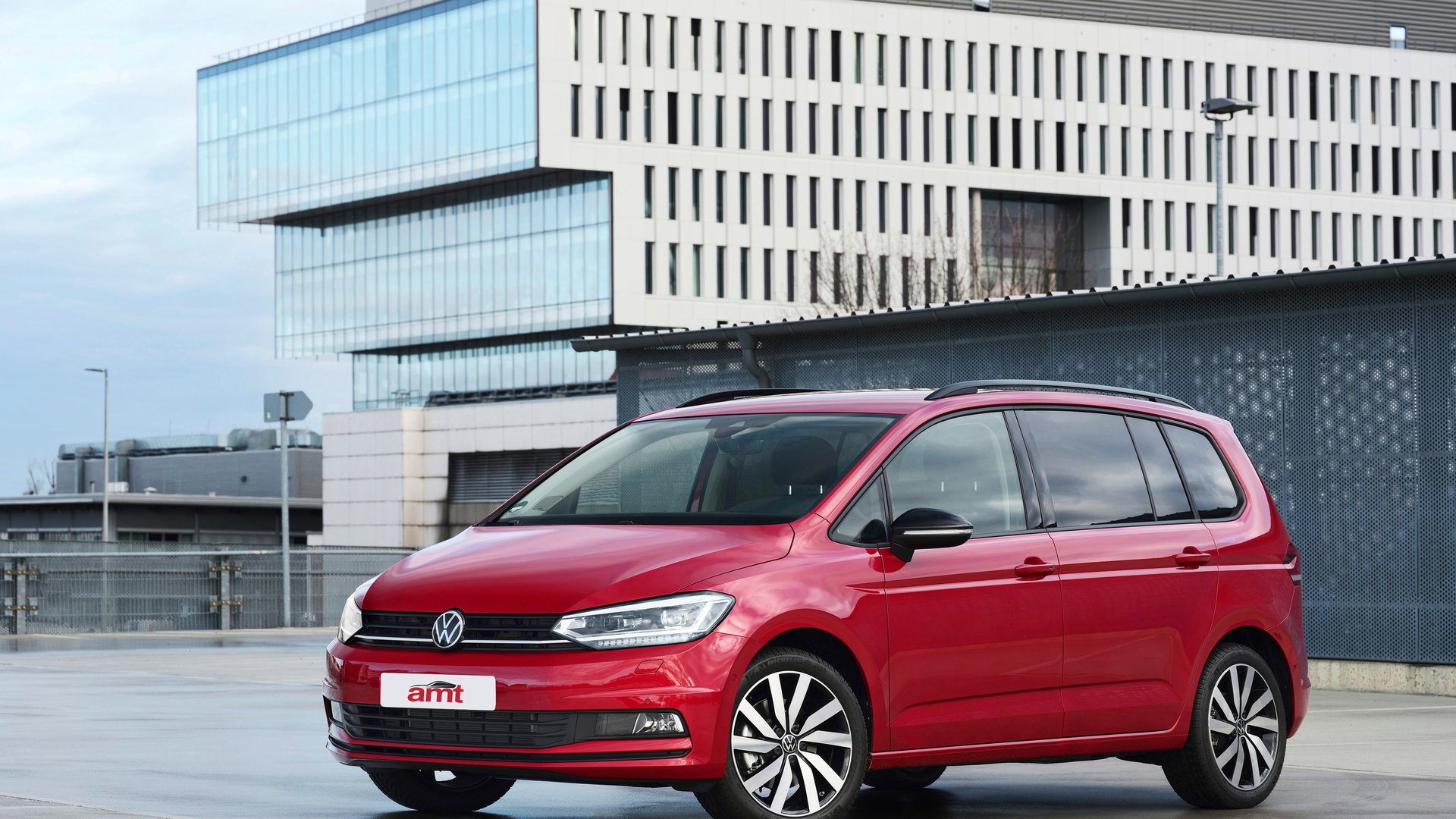 A red Volkswagen Touran, a practical and family-friendly 7 seater car, parked in a modern urban setting with sleek glass-fronted buildings in the background