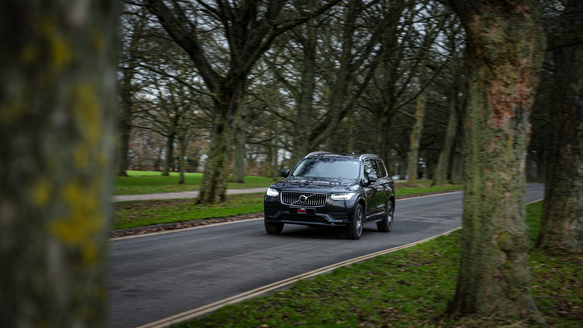 A black Volvo XC90, a luxury 7 seater car, driving through a tree-lined road in a serene park setting, highlighting its premium design and spaciousness