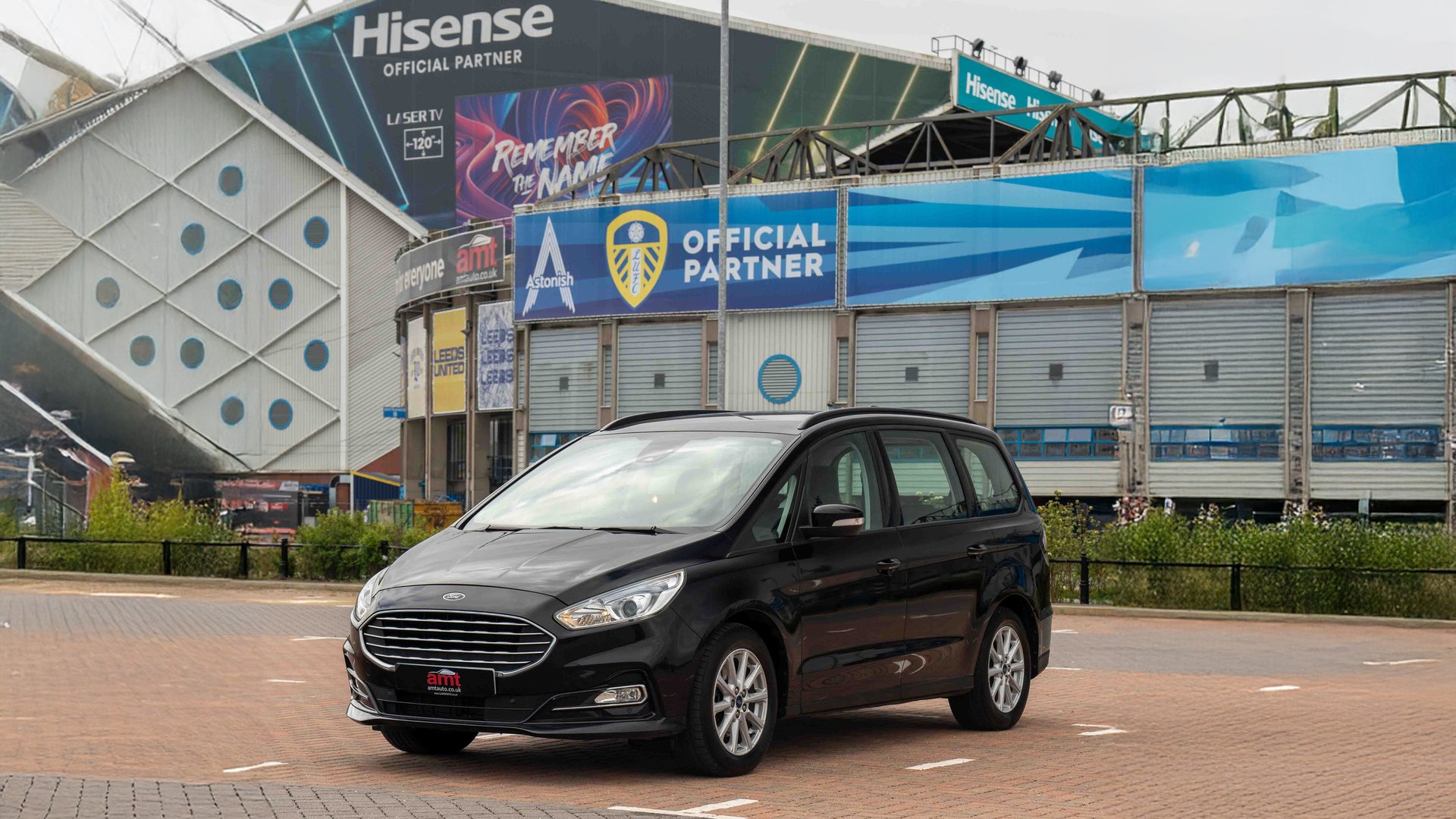 A black Ford Galaxy, a spacious and versatile 7 seater car, parked in front of the Leeds United stadium with vibrant sponsor banners and signage in the background