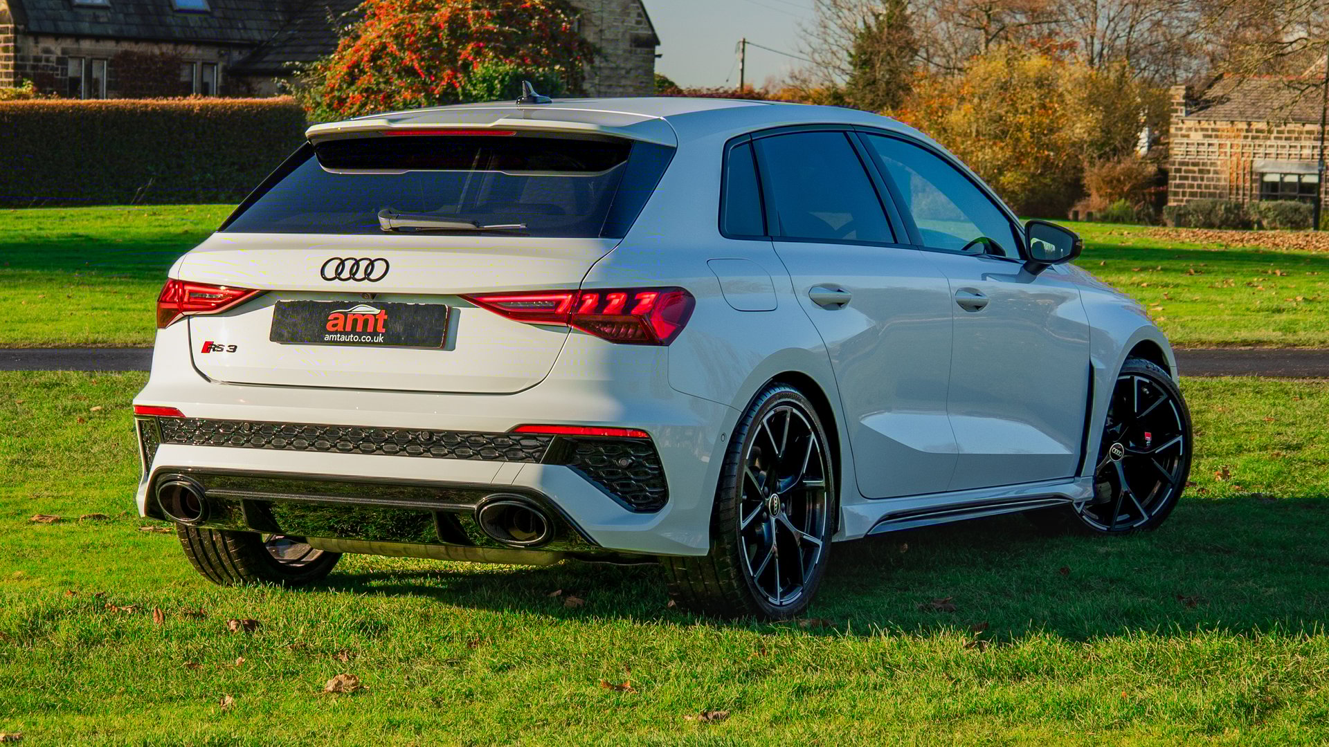 A rear view of the Audi RS3, highlighting its sporty tail lights, dual exhaust pipes, and stylish rear bumper with a rural setting in the background.