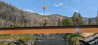 Ironworker guiding a steel bridge girder into place with a crane hook over a mountain river surrounded by forested hillsides
