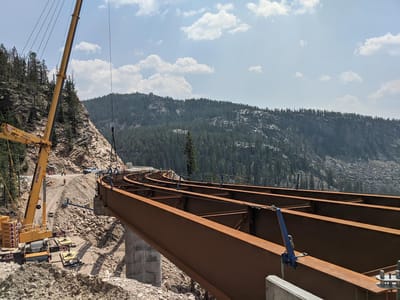 Steel arch bridge sections being erected by crane in a forested mountain valley at the Badger Creek project