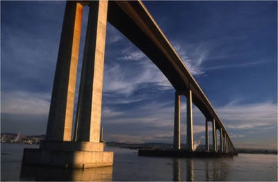 Underside of the Antioch Bridge at dusk showing tall concrete piers illuminated by warm light and reflected in calm water