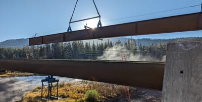 Crane lifting a steel plate girder into position during bridge construction over a forested river valley