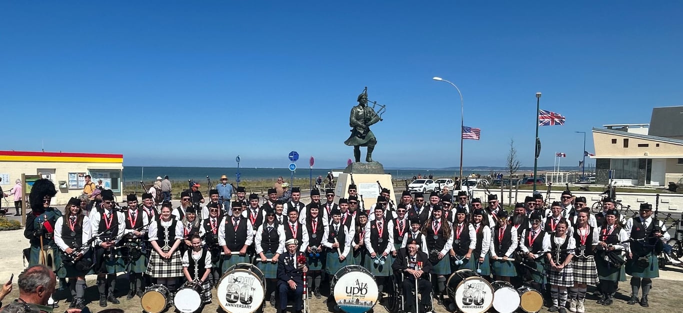 Utah Pipe Band in full highland dress posed in front of a piper statue memorial with flags