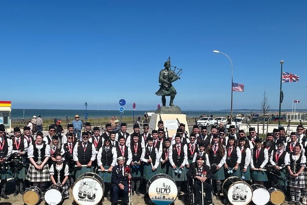 Utah Pipe Band in full highland dress posed in front of a piper statue memorial with flags