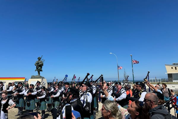 Large group of bagpipers performing in front of a soldier statue with American and British flags