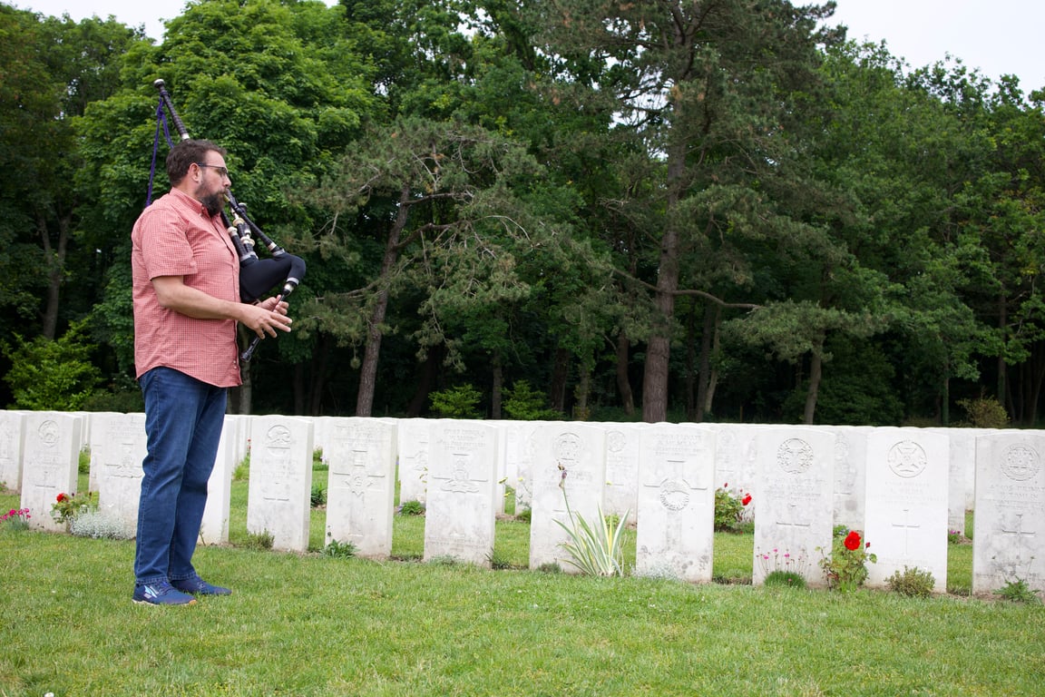 Man playing bagpipes among white headstones at a Commonwealth war cemetery surrounded by trees