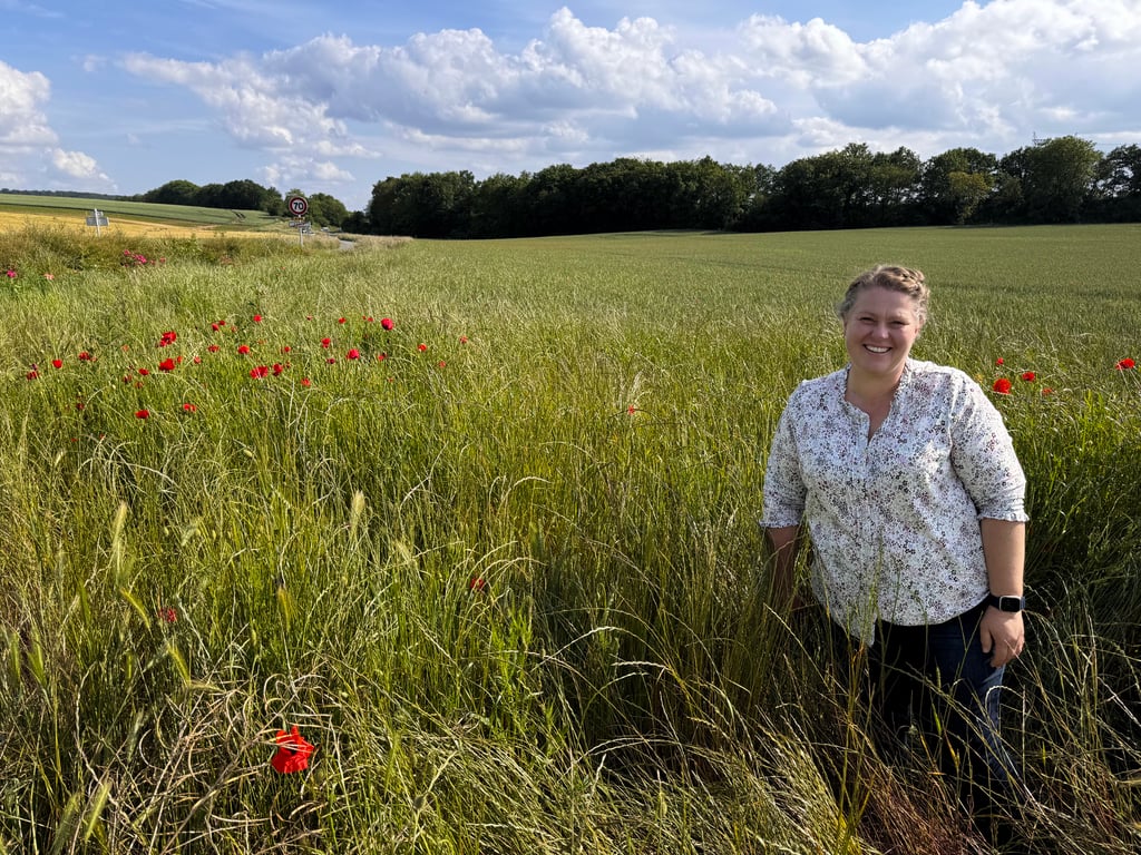 Woman standing in a lush green field dotted with red poppies under a blue sky in the French countryside