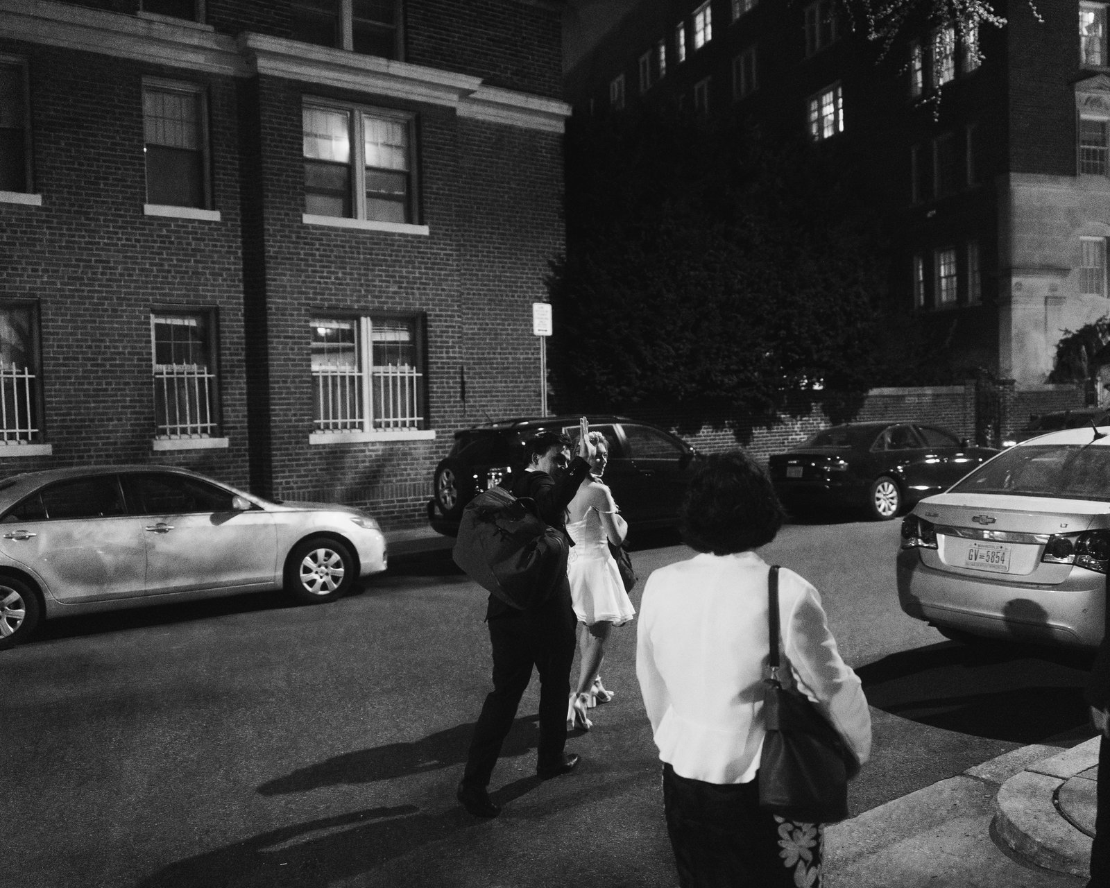 Black and white, the bride and groom walking down the street past parked cars at night