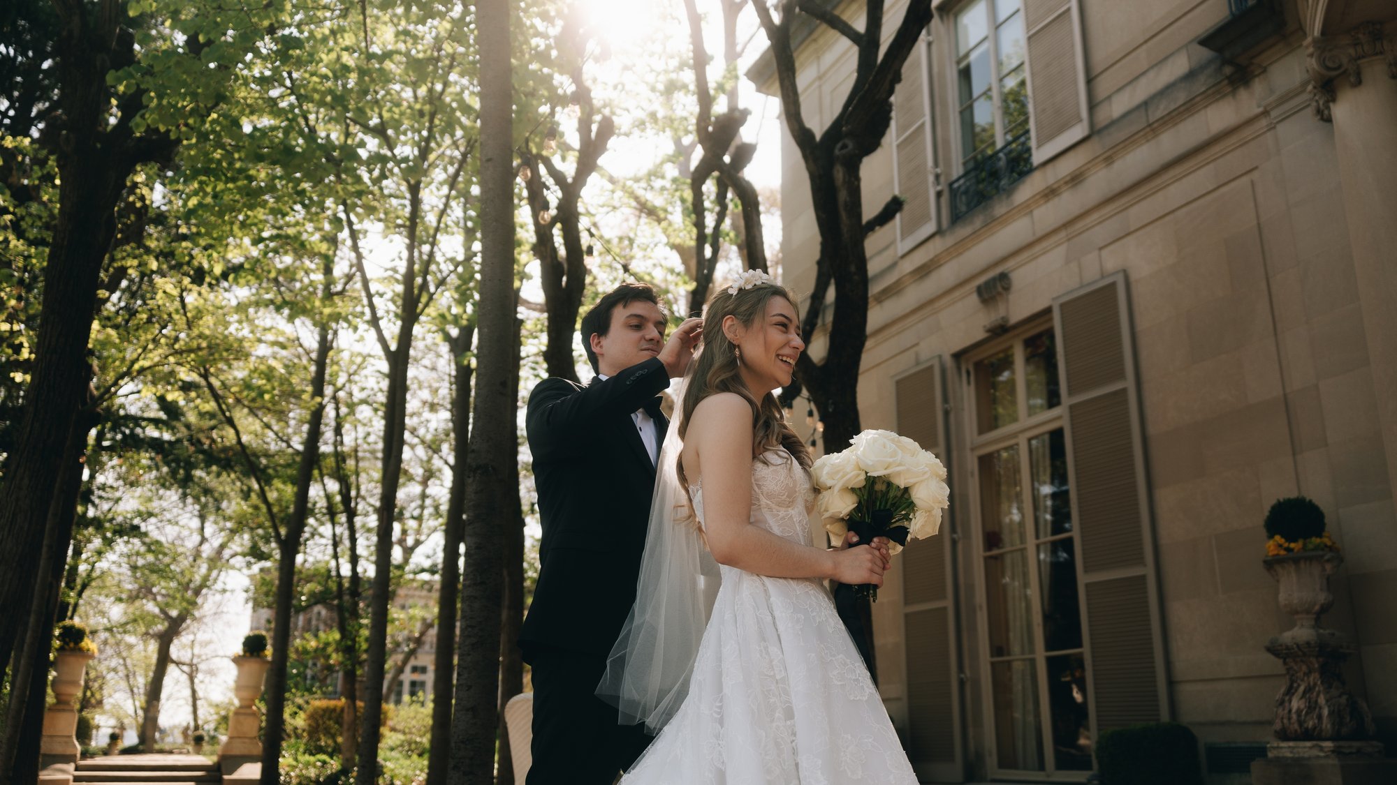 The bride and groom under bare spring trees beside the stone house