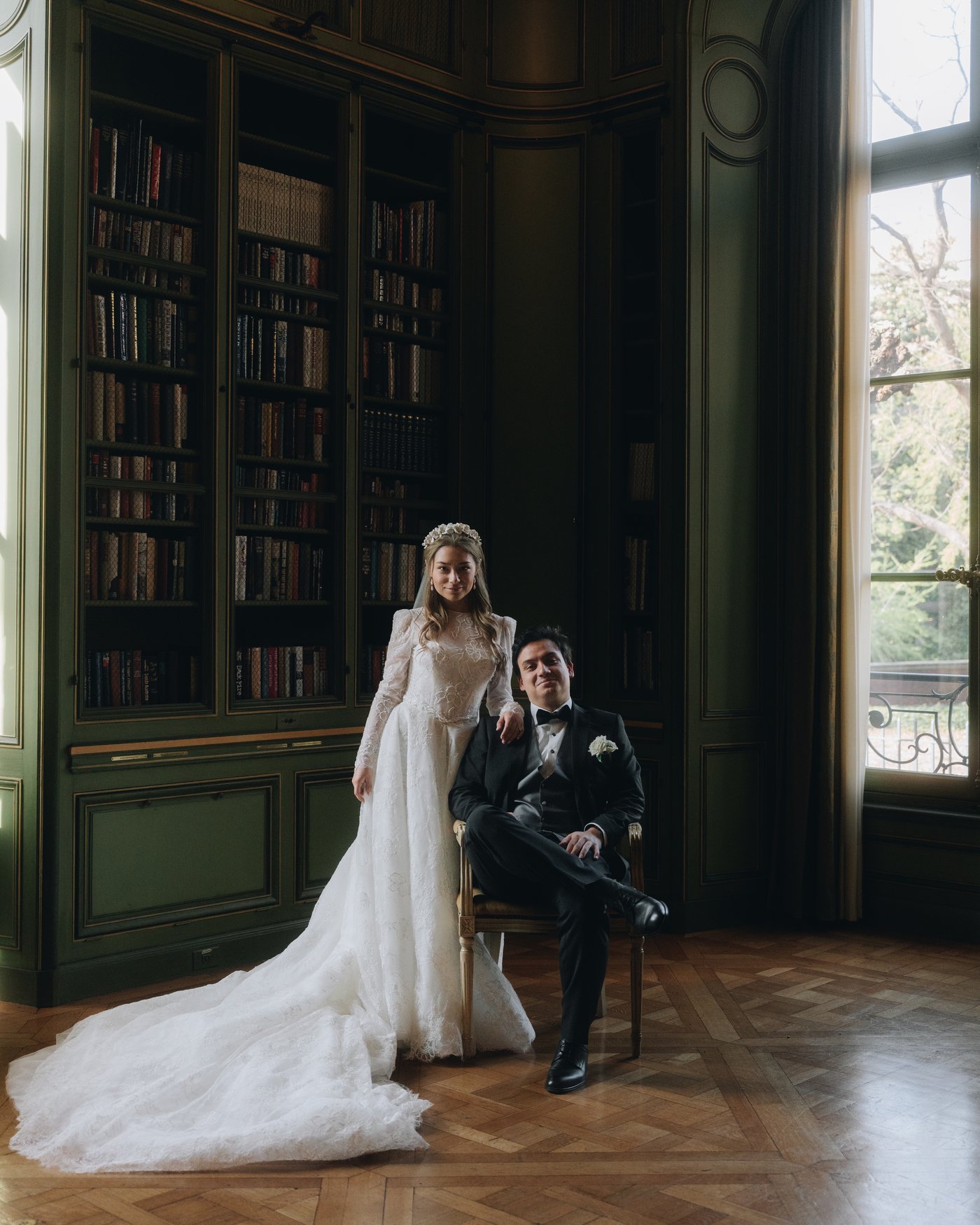 The bride seated in the library with a guest, bookshelves and tall windows behind them