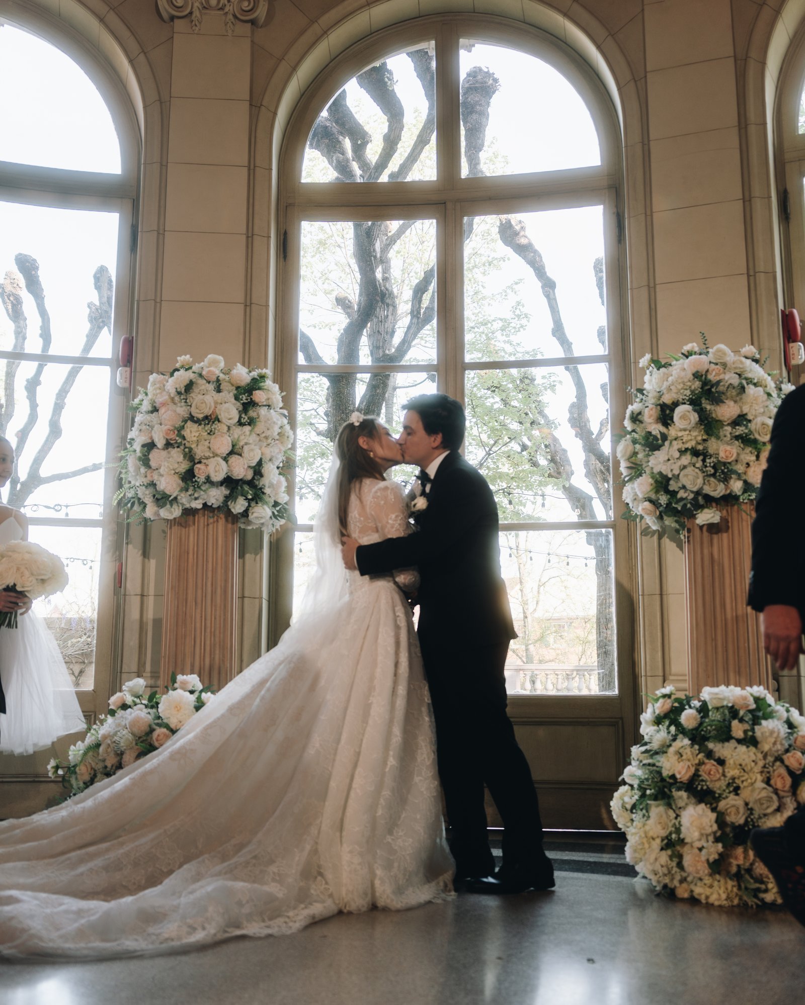 The bride and groom facing each other between two tall white floral urns beneath the arched windows