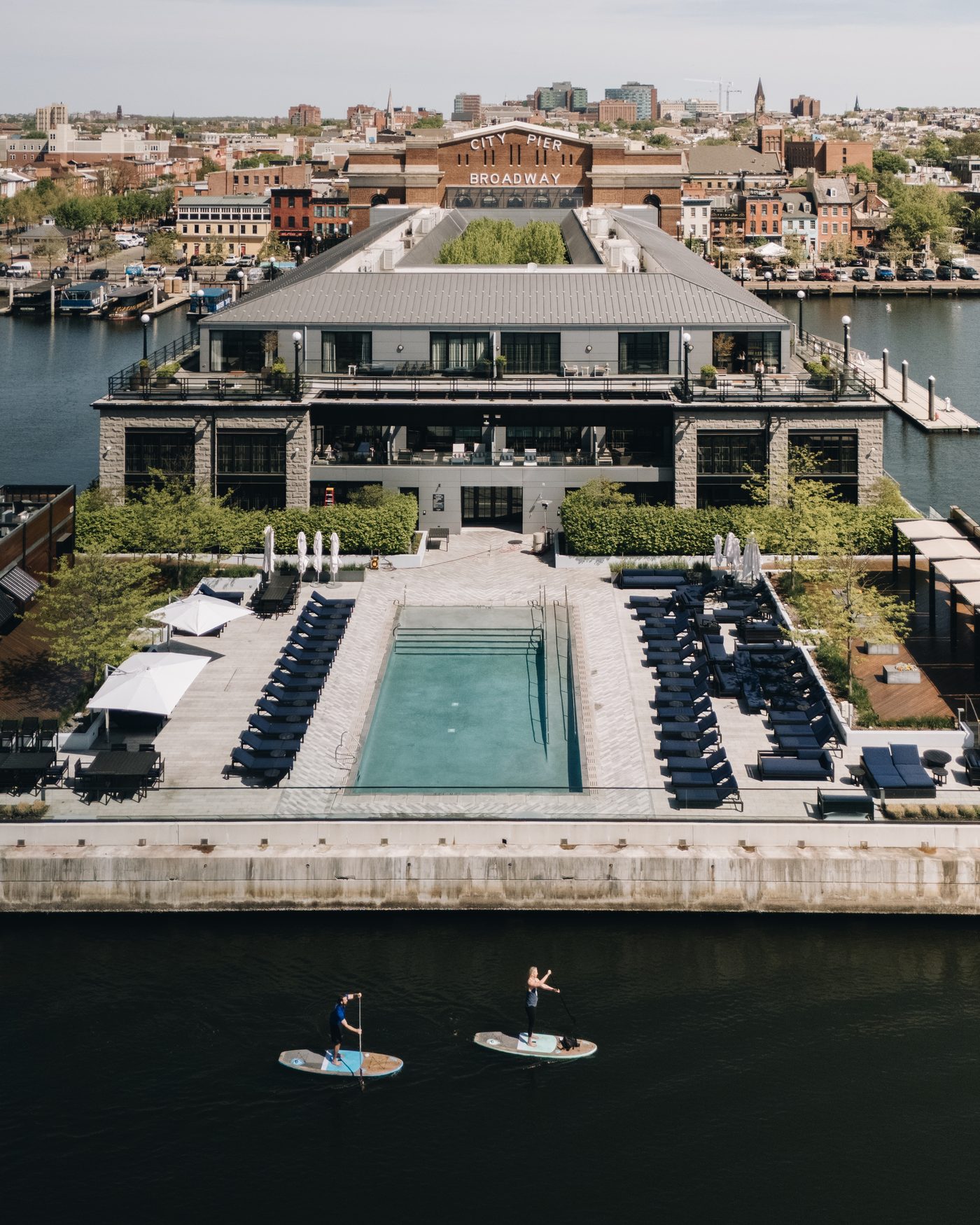 From out on the water, the low waterfront building with its pool and rows of loungers, city rooftops behind, two paddleboarders crossing the foreground