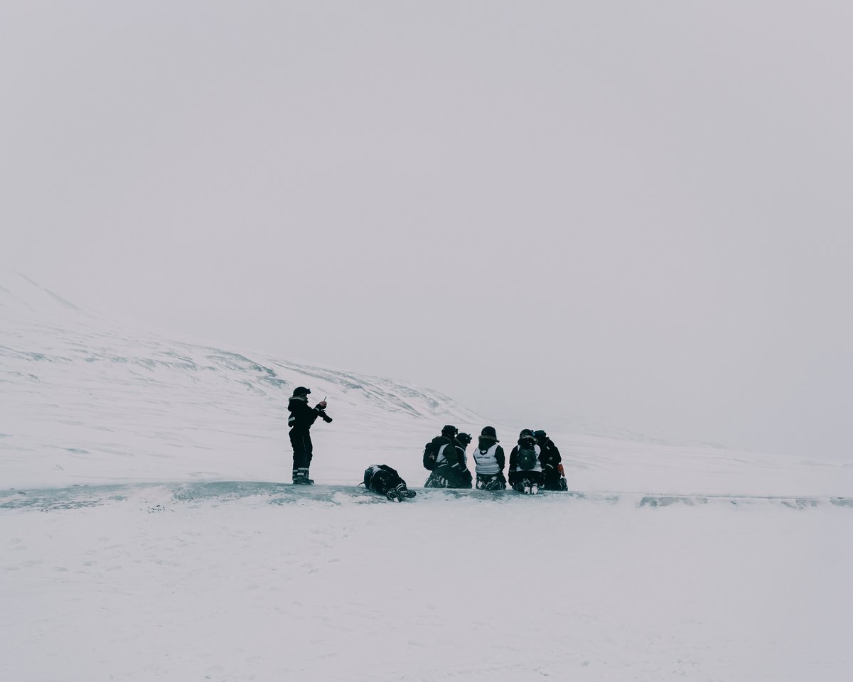 A photographer standing apart on a wide snowfield in flat white light, the group of guests kneeling together on the ice ahead of him