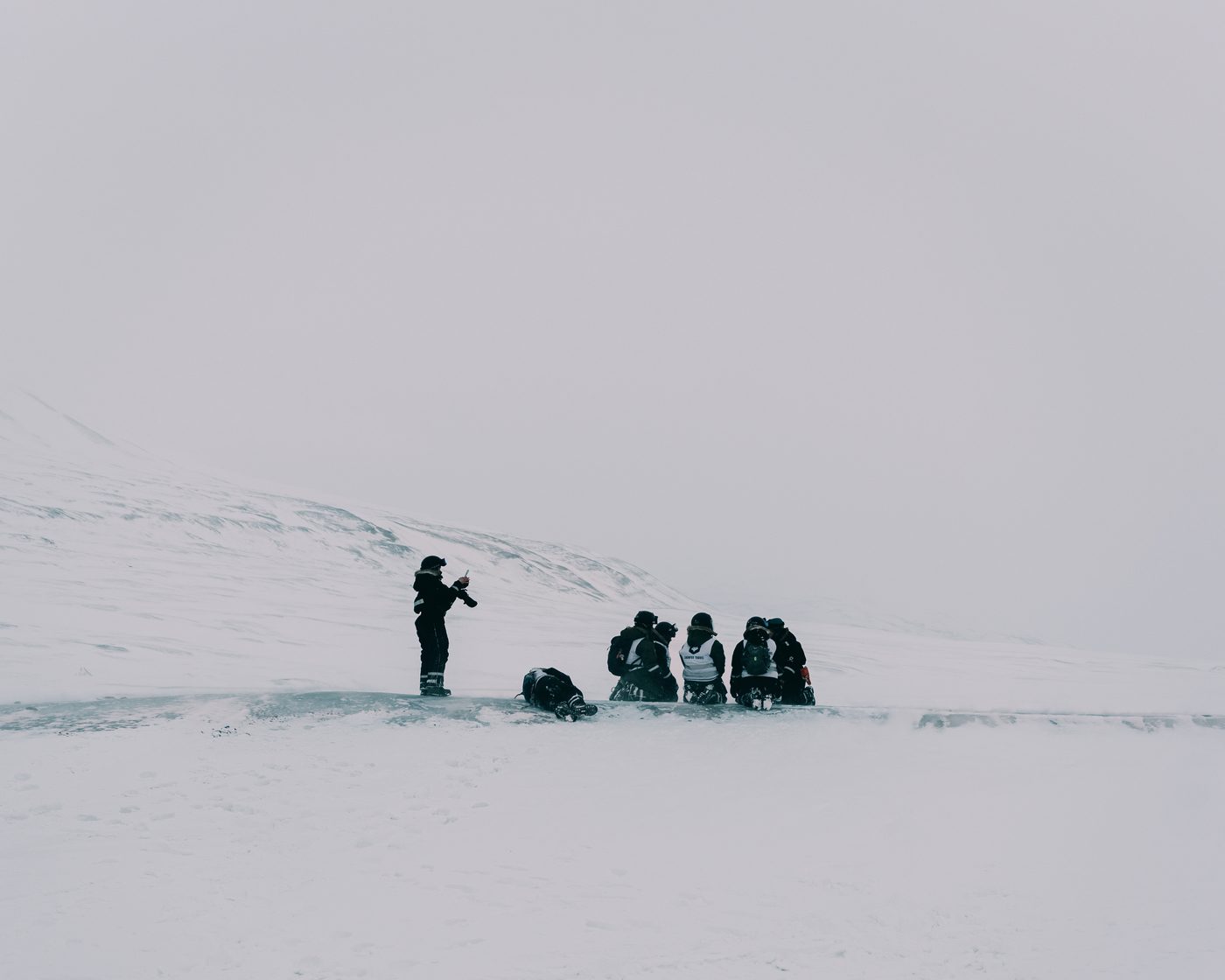 A photographer standing apart on a wide snowfield in flat white light, the group of guests kneeling together on the ice ahead of him