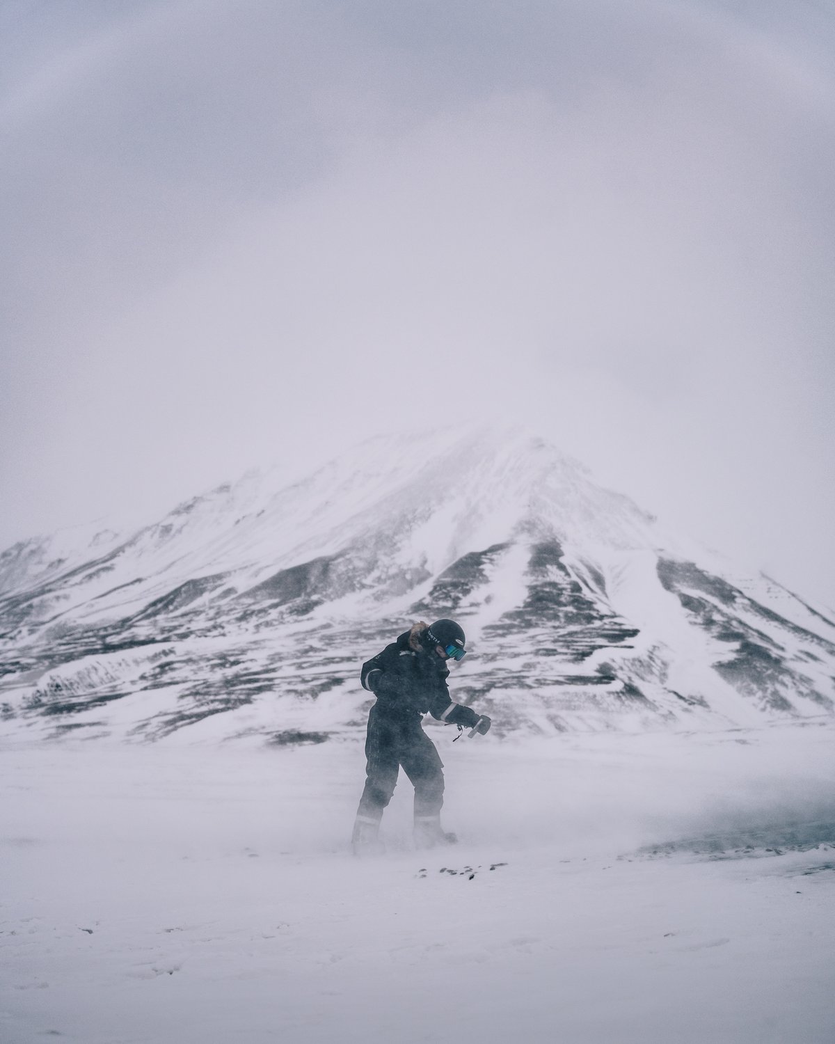 A guide crossing a frozen plain in blowing spindrift, a striped snow mountain rising into cloud behind