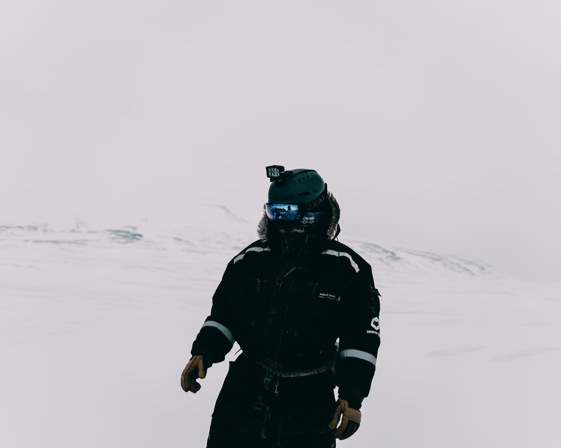 A guest in a black expedition suit and helmet alone on an empty white plain, the rest of the group reflected small in the mirrored goggles