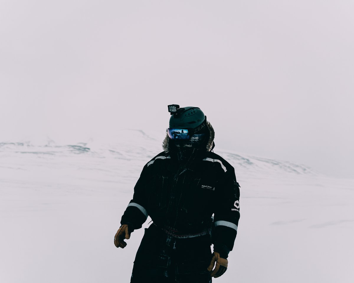 A guest in a black expedition suit and helmet alone on an empty white plain, the rest of the group reflected small in the mirrored goggles