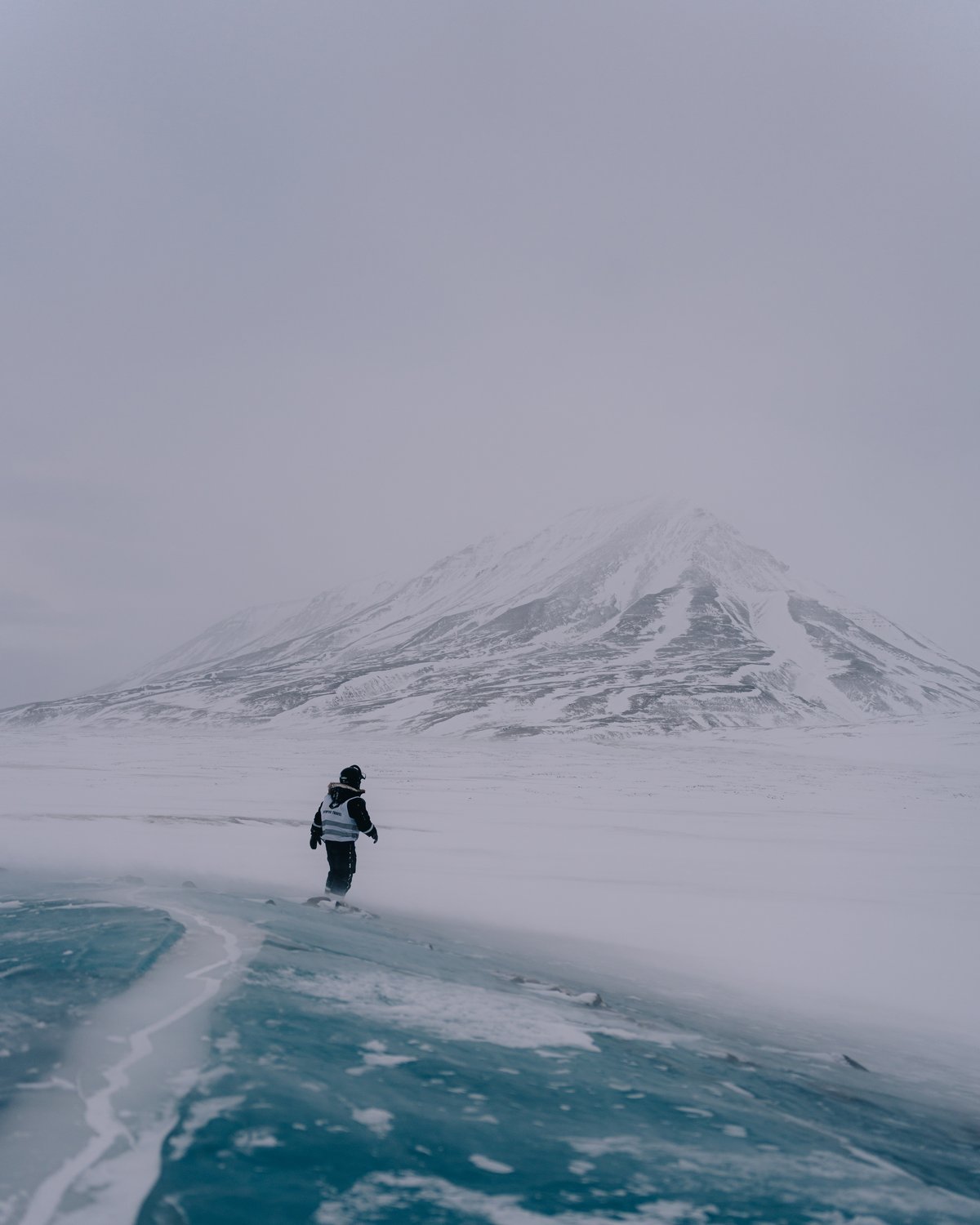 A lone figure in a high-visibility vest standing on bare turquoise lake ice, drifting snow crossing the foot of a snow mountain behind