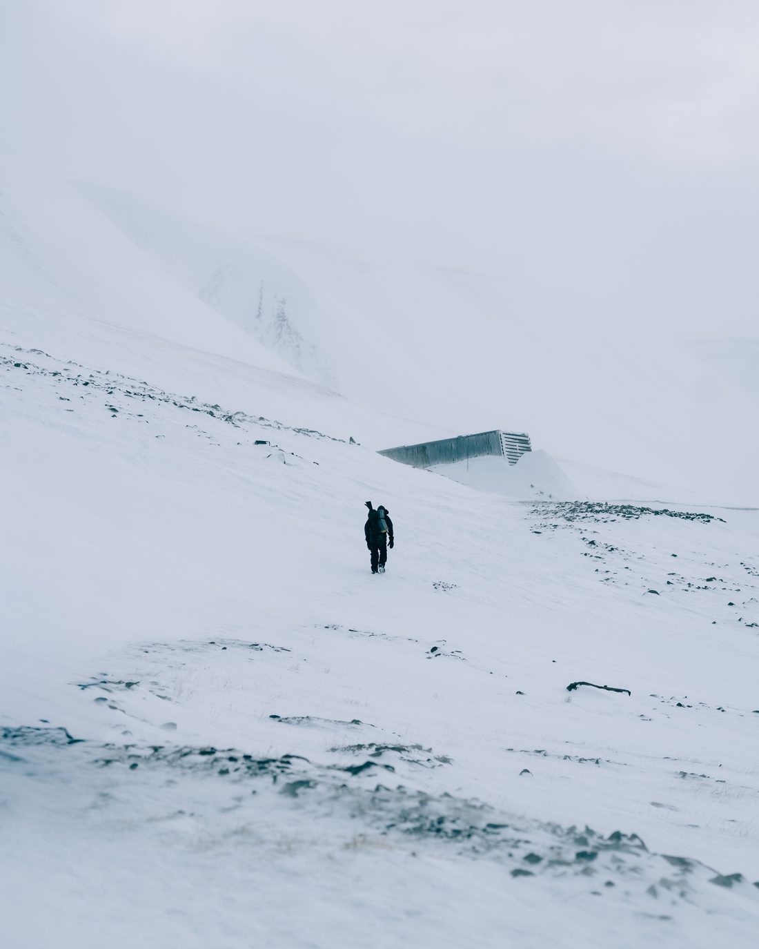A single walker crossing a wide snow slope toward the low dark entrance building set into the ridge above