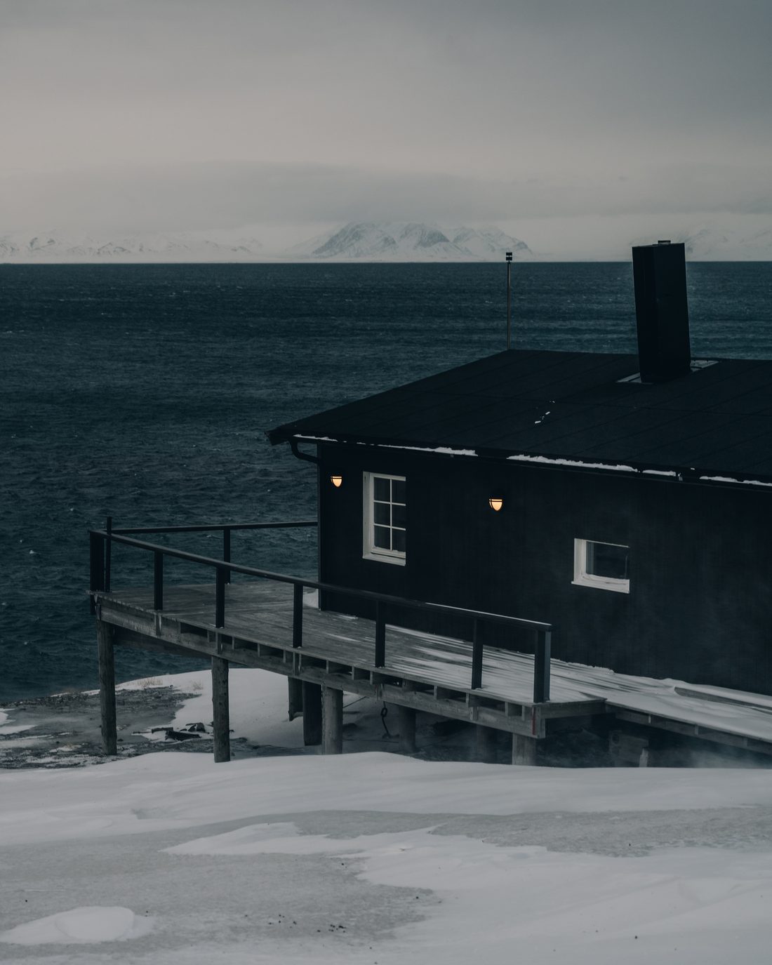 A black timber cabin on stilts at the water edge, two warm wall lamps burning against the dark sea and a snow range on the far shore