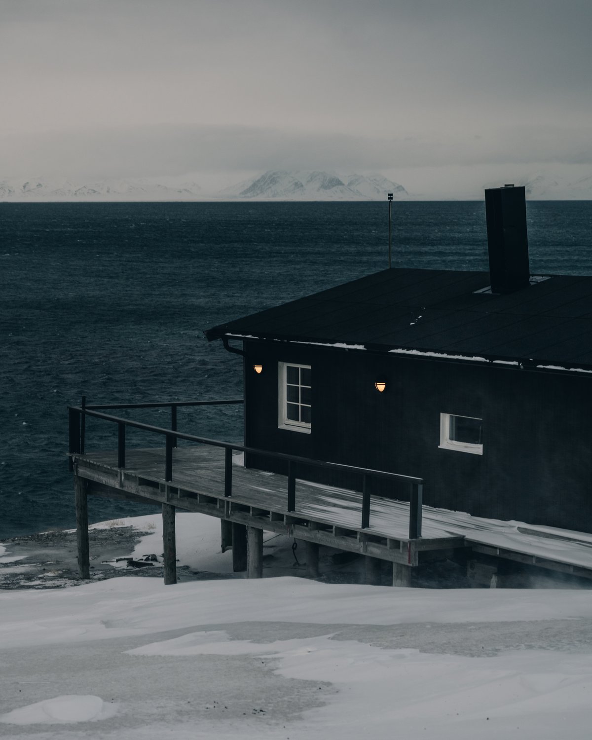 A black timber cabin on stilts at the water edge, two warm wall lamps burning against the dark sea and a snow range on the far shore